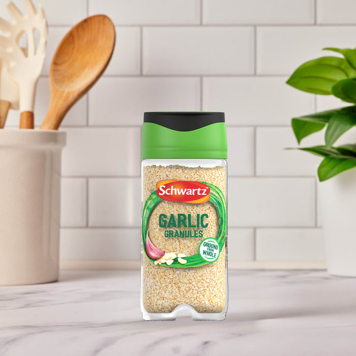 A clear jar of Schwartz Garlic Granules 50G sits on a white marble kitchen counter, with a utensil holder and a green plant blurred in the background.