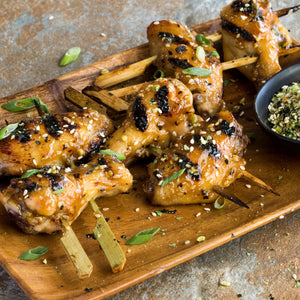 Grilled chicken wings on skewers are coated in a savory glaze, which could be made using Schwartz Garlic Granules 50G, and served on a wooden platter with a side of seasoning.