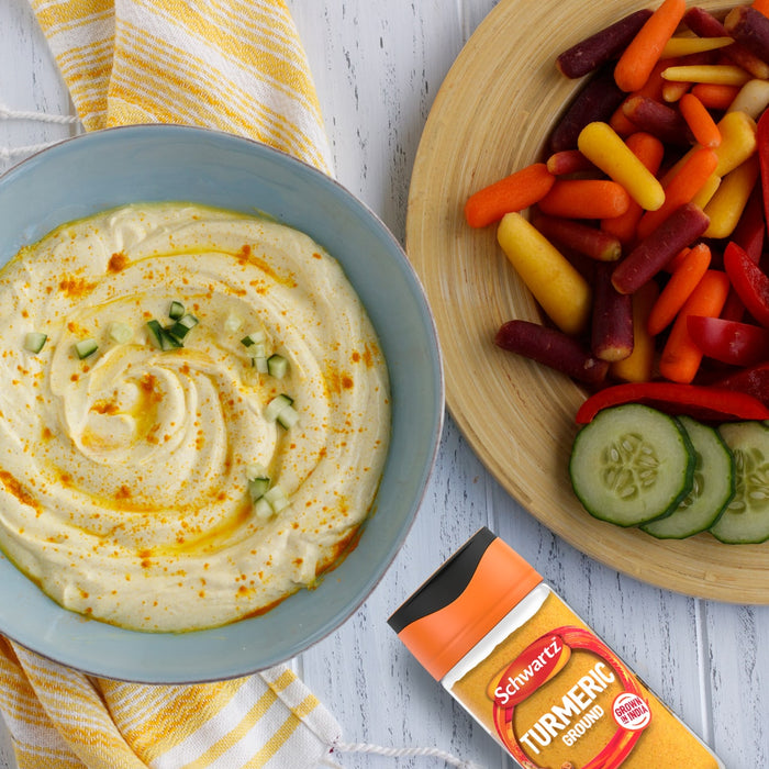 A bowl of creamy hummus is garnished with chopped cucumber and a sprinkle from a jar of Schwartz Turmeric Ground 37G, served alongside a platter of fresh vegetables for dipping.
