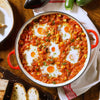 A vibrant shakshuka with eggs poached in a tomato and vegetable stew, a dish that can be beautifully flavored with Schwartz Turmeric Ground 37G, is presented in a red pan on a wooden table.