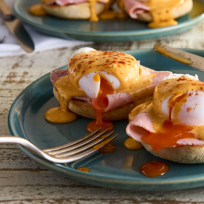 A delicious plate of Eggs Benedict is shown, with the runny yolk of a poached egg spilling out over ham and an English muffin, all topped with hollandaise sauce and a sprinkle of Schwartz Paprika 40G.