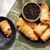 Crispy, golden-brown egg rolls filled with chicken and vegetables seasoned with Schwartz Onion Granules 30G are served on black plates with a spicy dipping sauce.