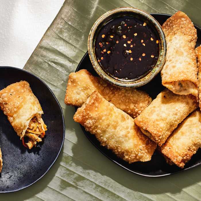 Crispy, golden-brown egg rolls filled with chicken and vegetables seasoned with Schwartz Onion Granules 30G are served on black plates with a spicy dipping sauce.