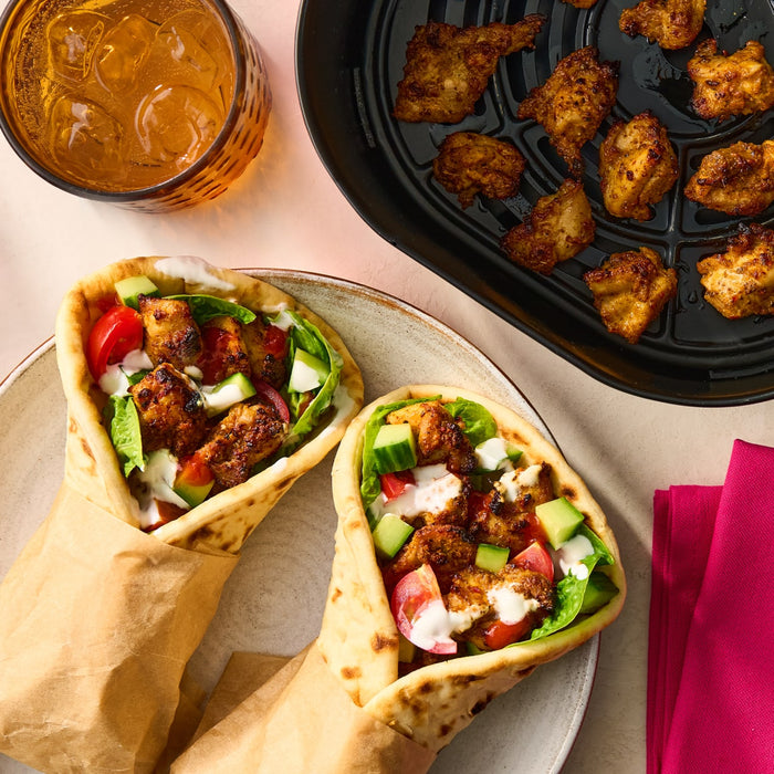 Two pita wraps filled with fresh vegetables and chicken seasoned with Schwartz Af Turki Keb Seas are served on a plate next to an air fryer basket and a glass of iced tea.