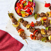 Grilled beef and vegetable skewers seasoned with Schwartz Af Turki Keb Seas are served alongside a vibrant tomato and onion salad on a marble surface.