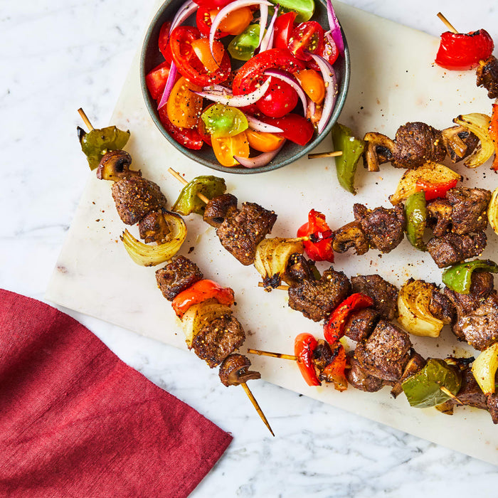Grilled beef and vegetable skewers seasoned with Schwartz Af Turki Keb Seas are served alongside a vibrant tomato and onion salad on a marble surface.