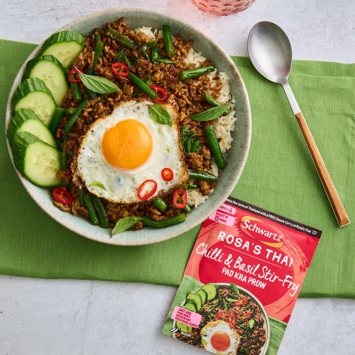 A finished dish of Thai Pad Kra Pao, featuring minced pork, green beans, and a fried egg on rice, is displayed in a bowl next to the Schwartz Sa Rt Chili Basil Stir Fry X12 seasoning packet used to prepare it.