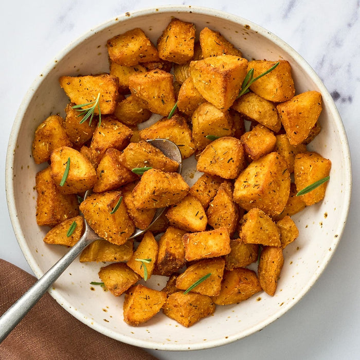 A bowl of crispy, golden-brown roasted potatoes made with Af Bvp Sch Potatoes Seas seasoning is garnished with fresh rosemary and served with a spoon.