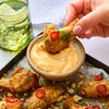 A hand dips a crispy chicken wing seasoned with Jr Thai Sweet Chil Seas X6 into a creamy dipping sauce bowl surrounded by more wings garnished with red chili slices and crushed peanuts.