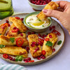 A hand dips a golden-brown halloumi fry seasoned with Jr Hot Honey Seas X6 into a creamy yogurt dip garnished with olive oil and herbs on a plate with pomegranate seeds and chili slices.