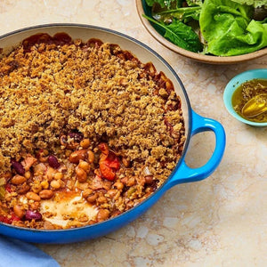 A hearty bean crumble bake is served alongside a fresh green salad and a small bowl of oil infused with Schwartz Italian Herb Seasoning 11G.