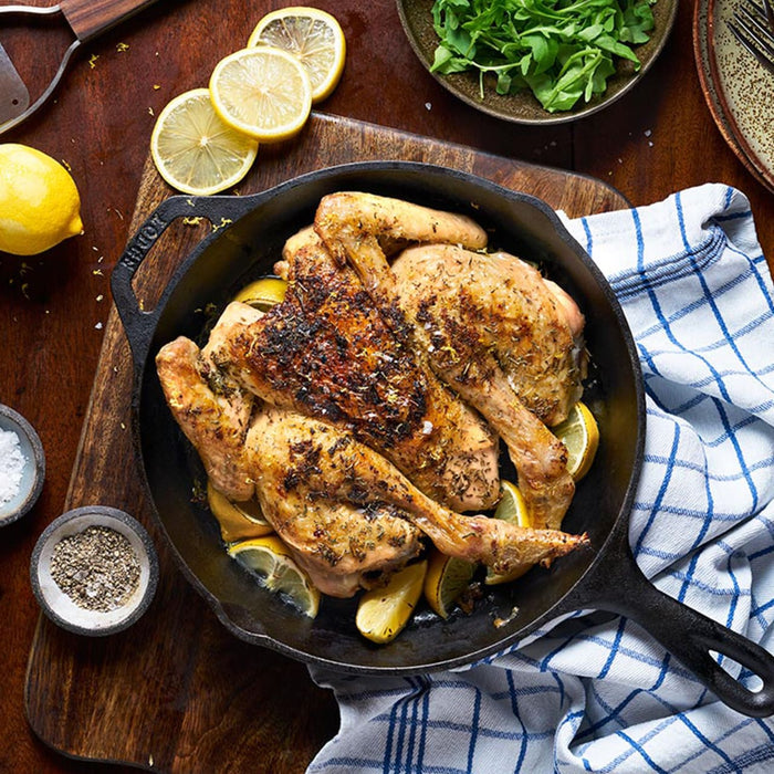 A spatchcocked chicken, beautifully roasted with lemon and seasoned with Schwartz Thyme 11 G, is presented in a cast-iron skillet on a wooden table.