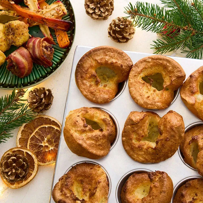 A festive Christmas dinner is laid out, featuring golden Yorkshire puddings in a tin and a plate of roasted vegetables and pigs in a blanket seasoned with Schwartz Thyme 11 G.