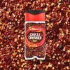 A clear jar of Schwartz Crushed Chillies 29G is centered on a vibrant background of the same red crushed chilli flakes.