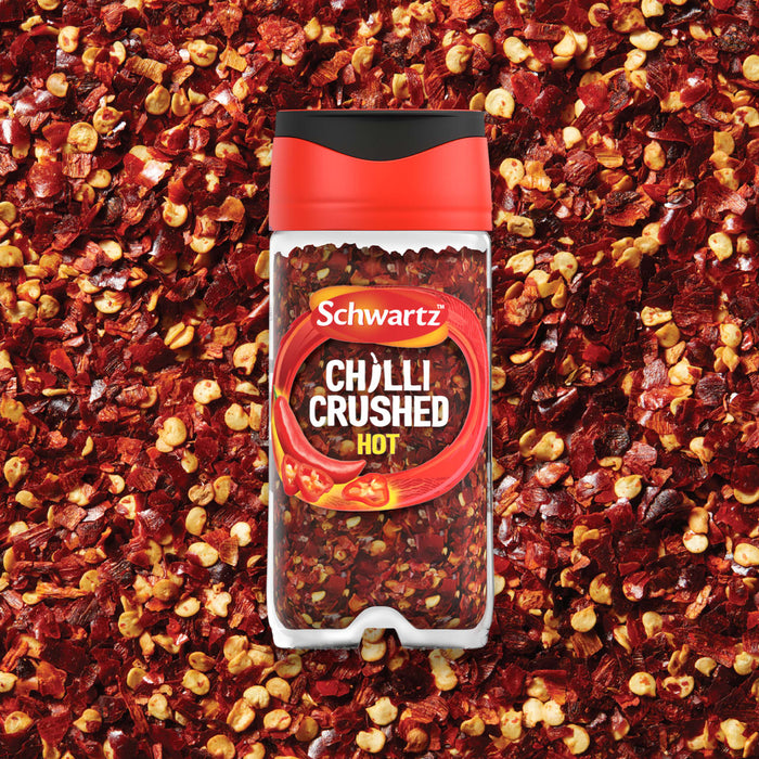 A clear jar of Schwartz Crushed Chillies 29G is centered on a vibrant background of the same red crushed chilli flakes.