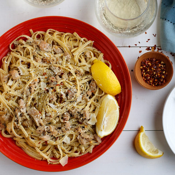 A red plate of linguine with clams is served with lemon wedges, a glass of white wine, and a small bowl of Schwartz Crushed Chillies 29G for sprinkling on top.