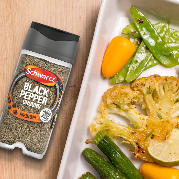 A jar of Schwartz Ground Black Pepper, 33G, sits on a wooden surface next to a white dish of roasted vegetables, suggesting a flavorful seasoning option.