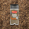 A clear plastic container of Schwartz Ground Black Pepper, 33G, is shown centered on a bed of coarse ground black pepper.