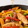 A cast iron skillet filled with sizzling chicken strips and colorful bell peppers being seasoned with Schwartz Fajita Seasoning 46G.