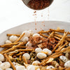 A savory snack mix featuring pretzel sticks and puffed lotus seeds is being dusted with Schwartz Cajun Seasoning 44G for a bold and spicy flavor.