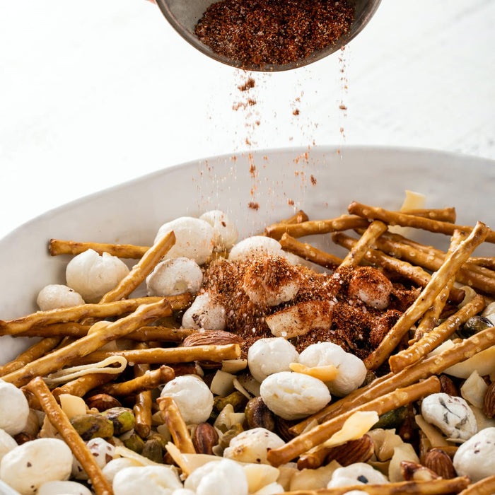 A savory snack mix featuring pretzel sticks and puffed lotus seeds is being dusted with Schwartz Cajun Seasoning 44G for a bold and spicy flavor.