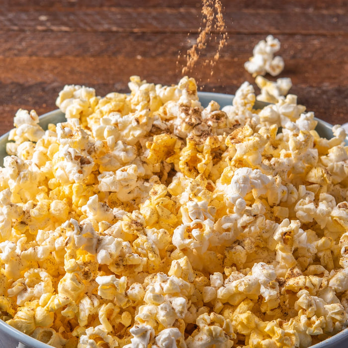 A bowl of popcorn is being seasoned with a sprinkle of Schwartz Garam Masala, 30G for a flavorful snack.