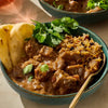 A delicious bowl of homemade lamb curry, made with the rich and aromatic flavors of Schwartz Garam Masala, 30G, is served with rice pilaf, naan bread, and a fresh cilantro garnish.