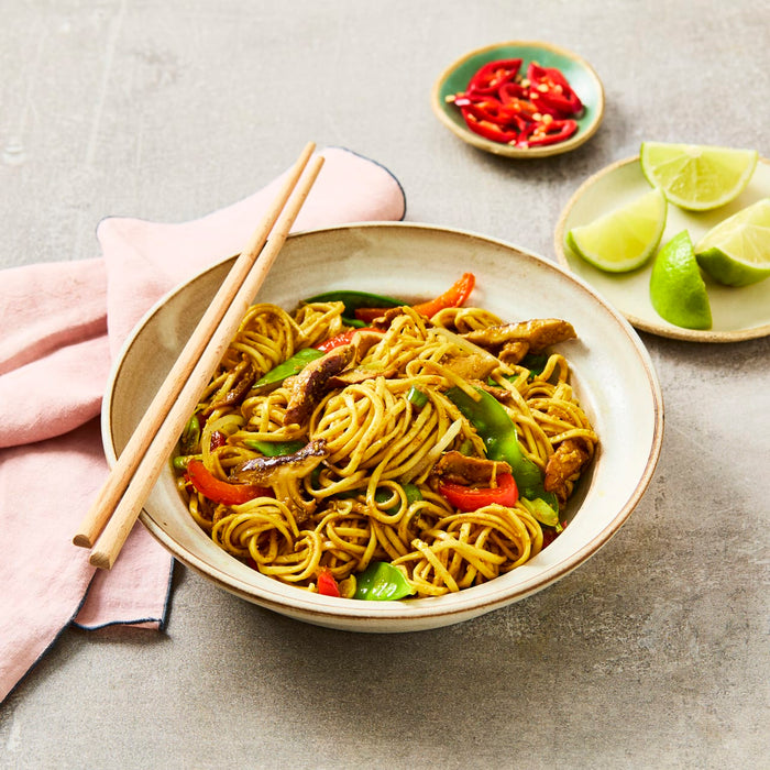 A rustic bowl filled with stir-fried noodles, chicken, and vegetables, served with chopsticks and a side of fresh lime and chili.