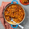 A vibrant blue cast iron pot holds a savory rice and meat pilaf, with a large spoon ready for serving and a portion already on a red plate.