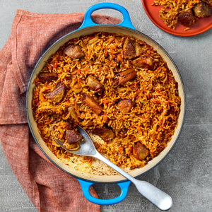 A vibrant blue cast iron pot holds a savory rice and meat pilaf, with a large spoon ready for serving and a portion already on a red plate.