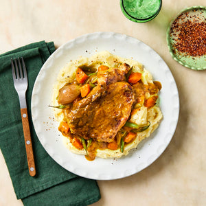 A delicious-looking pork chop smothered in a rich gravy is served on a bed of mashed potatoes with carrots and green beans, with a side of red pepper flakes for extra spice.