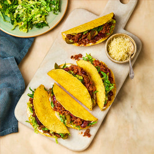 A top-down view of several crispy tacos filled with seasoned ground meat and beans, arranged on a white serving board with bowls of shredded lettuce and cheese nearby.