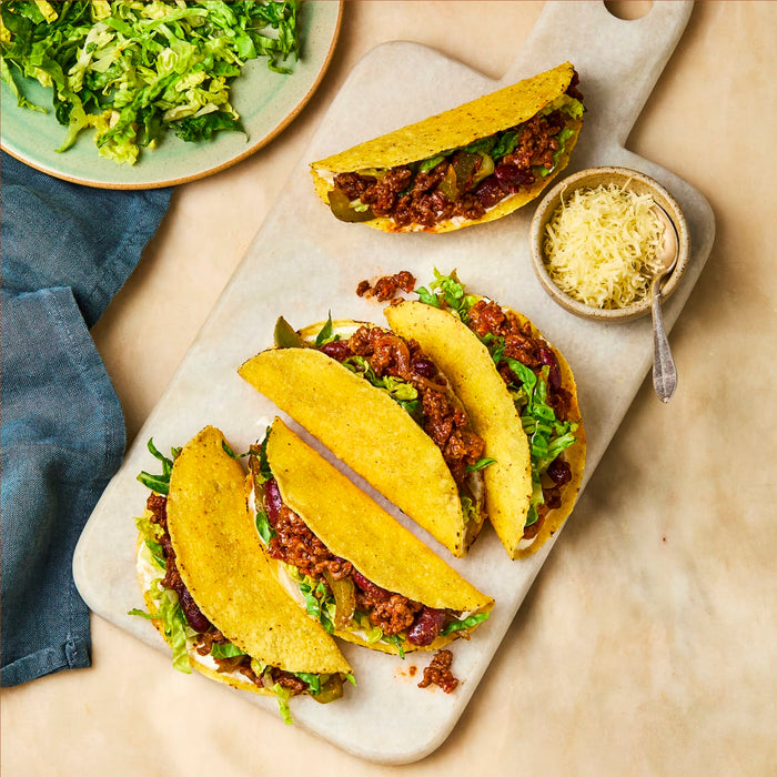 A top-down view of several crispy tacos filled with seasoned ground meat and beans, arranged on a white serving board with bowls of shredded lettuce and cheese nearby.
