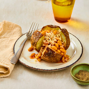 A baked potato is cut open on a white plate and filled with baked beans and shredded cheese, with a fork and a small bowl of pepper nearby.