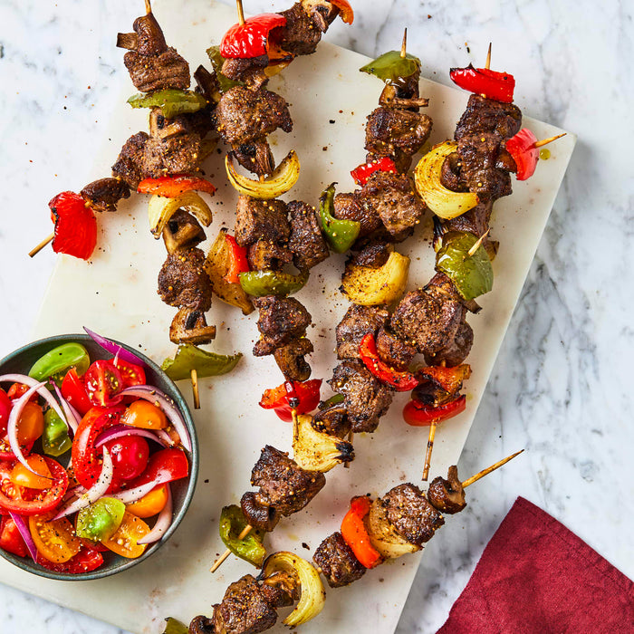 Grilled beef and vegetable skewers are arranged on a white marble platter next to a bowl of fresh tomato salad.