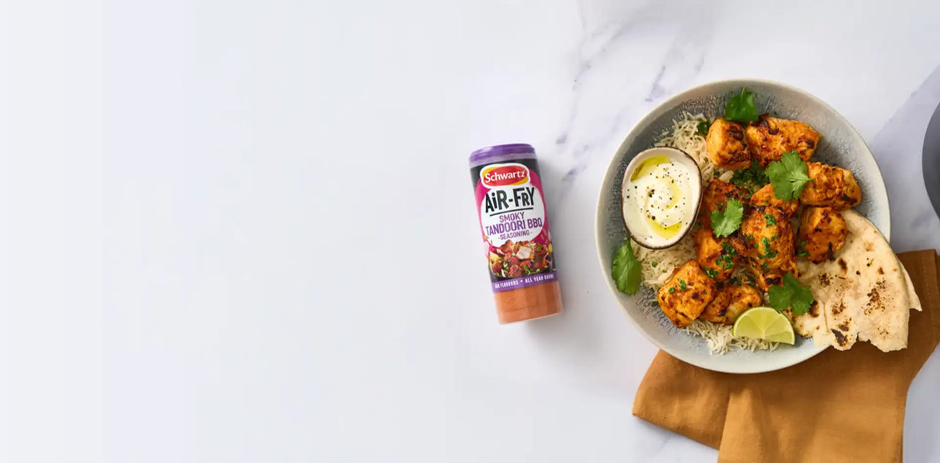 Bowl of chicken tikka masala with naan bread and a container of Schwartz air fry smoky tandoori bbq seasoning on a marble surface.