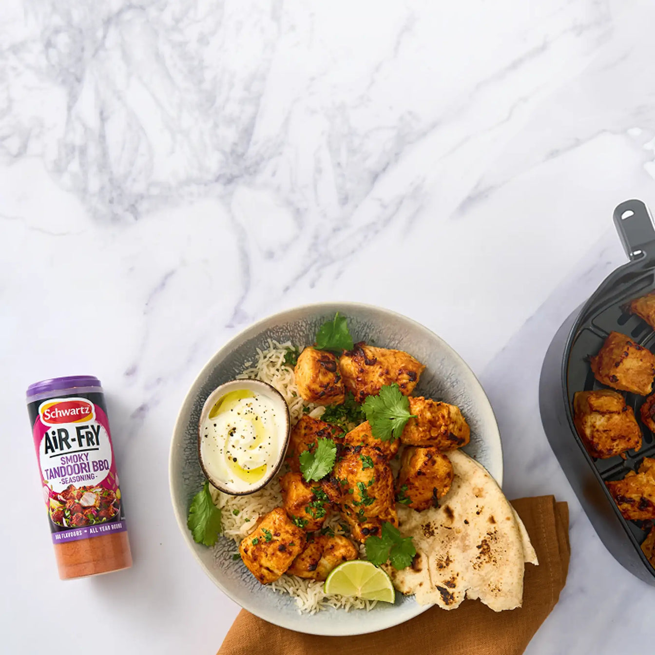 Bowl of chicken tikka masala with naan bread and a container of Schwartz air fry smoky tandoori bbq seasoning on a marble surface.