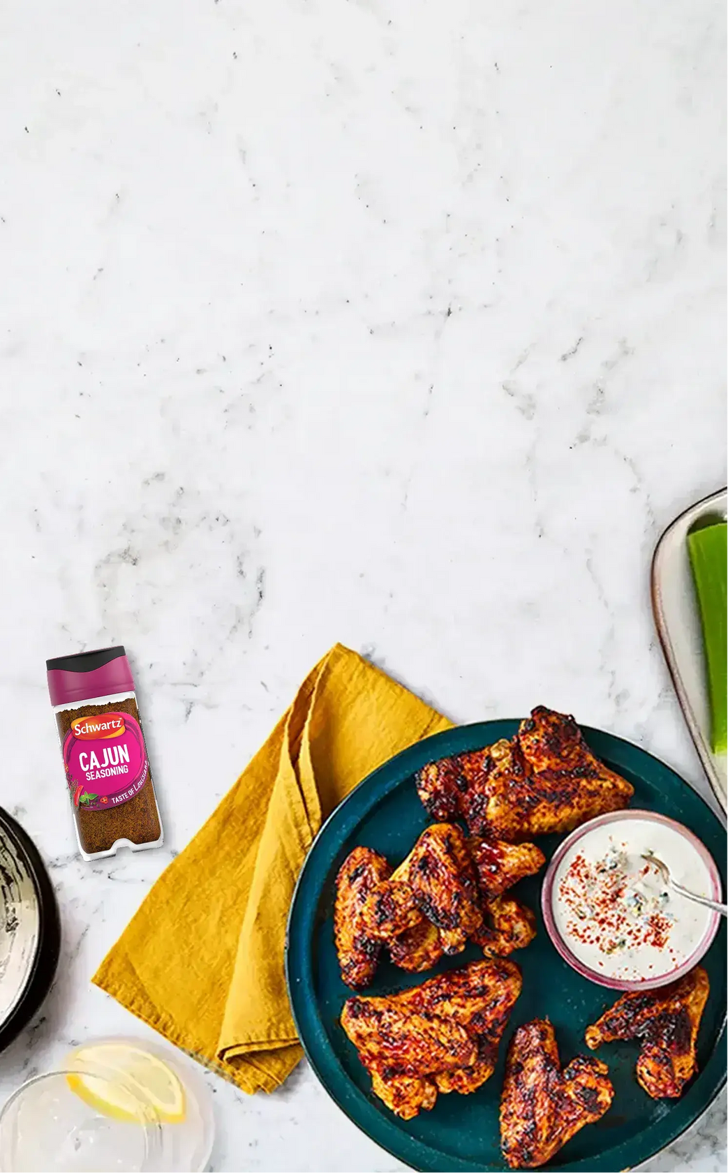 A styled overhead food scene on a white marble surface showing grilled chicken wings on a dark teal plate with a small bowl of white dipping sauce, a yellow napkin, and a spice jar labeled CAJUN Seasoning nearby.