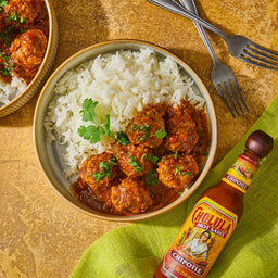Bowl of meatballs with rice and a bottle of Cholula hot sauce on a gold surface.