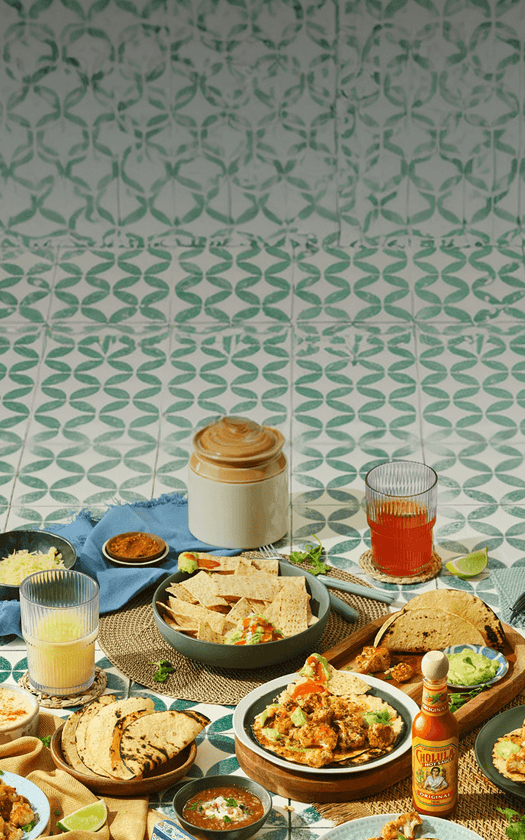 Diverse food items including tortillas, guacamole, and drinks on a patterned tablecloth.