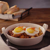 A savory breakfast hash with potatoes and meat is served in a white Barrel bowl, topped with two sunny-side-up eggs and a drizzle of hot sauce.