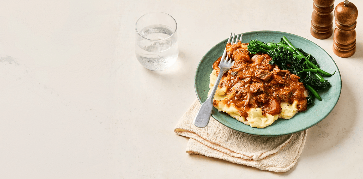 Plate of food with mashed potatoes, meat sauce, and greens on a light surface.