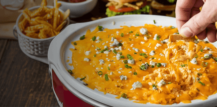 Buffalo chicken dip with a cracker being dipped into it, surrounded by chips and a sandwich.