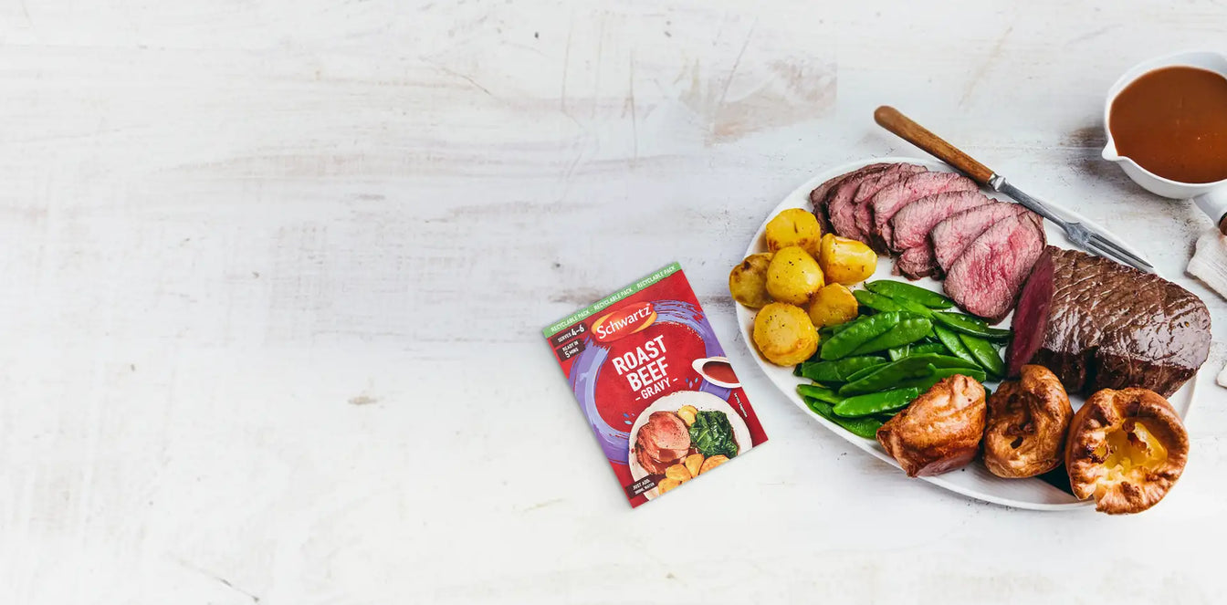 Plated roast beef with potatoes, peas, and Yorkshire pudding on a white surface with a packet of Schwartz roast beef seasoning.