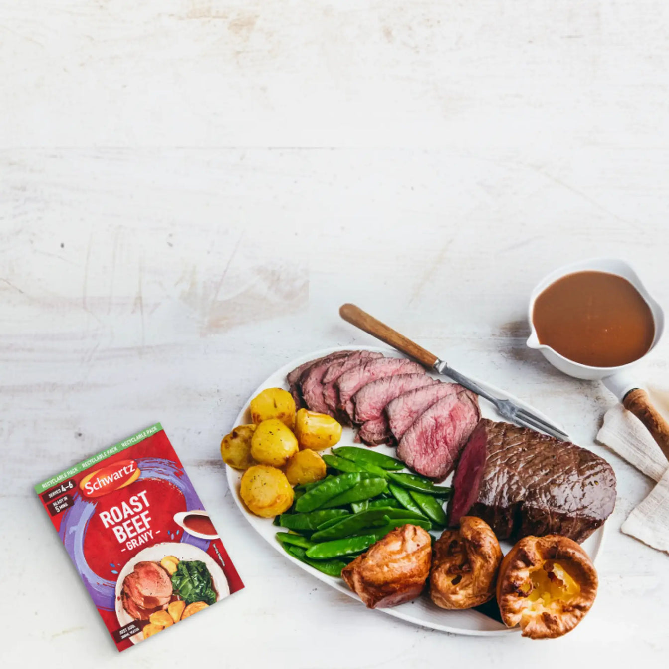 Plated roast beef with potatoes, peas, and Yorkshire pudding on a white surface with a packet of Schwartz roast beef seasoning.