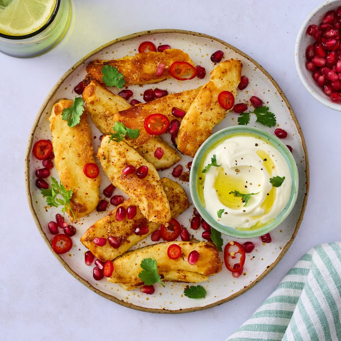 A plate of golden-brown fried halloumi sticks is garnished with pomegranate seeds, sliced red chili, and fresh cilantro, served with a creamy dipping sauce.