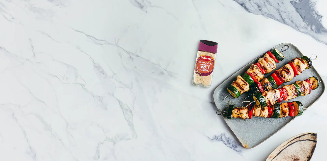Grilled vegetable skewers on a plate with a side salad and seasoning packet on a marble surface.