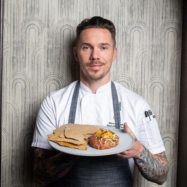Chef holding a plate with tartare and crackers against a patterned wall