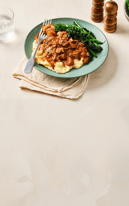 Plate of food with mashed potatoes, meat sauce, and greens on a beige surface.