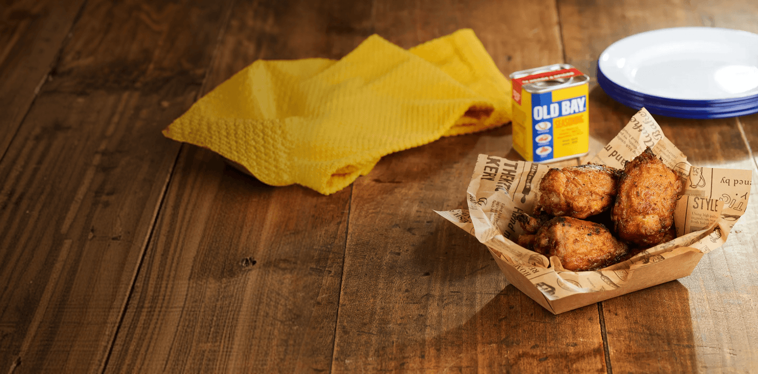 Fried chicken in a paper basket on a wooden table with a yellow cloth and Old Bay seasoning container.